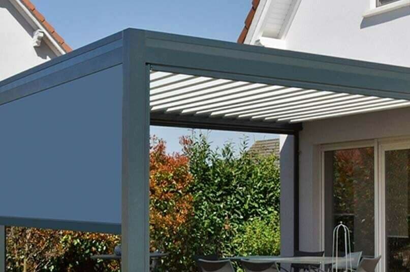 Pergola bioclimatique grise à lames orientables, fermée sur un côté par un store vertical bleu