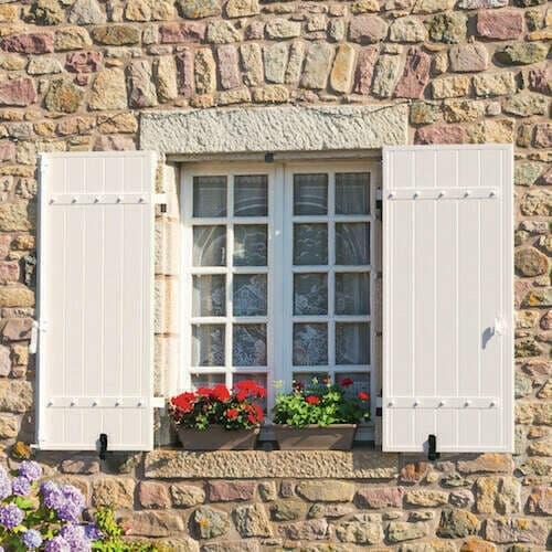 Volets battants en PVC blancs et fenêtre à petits bois fleurie sur une façade en pierre
